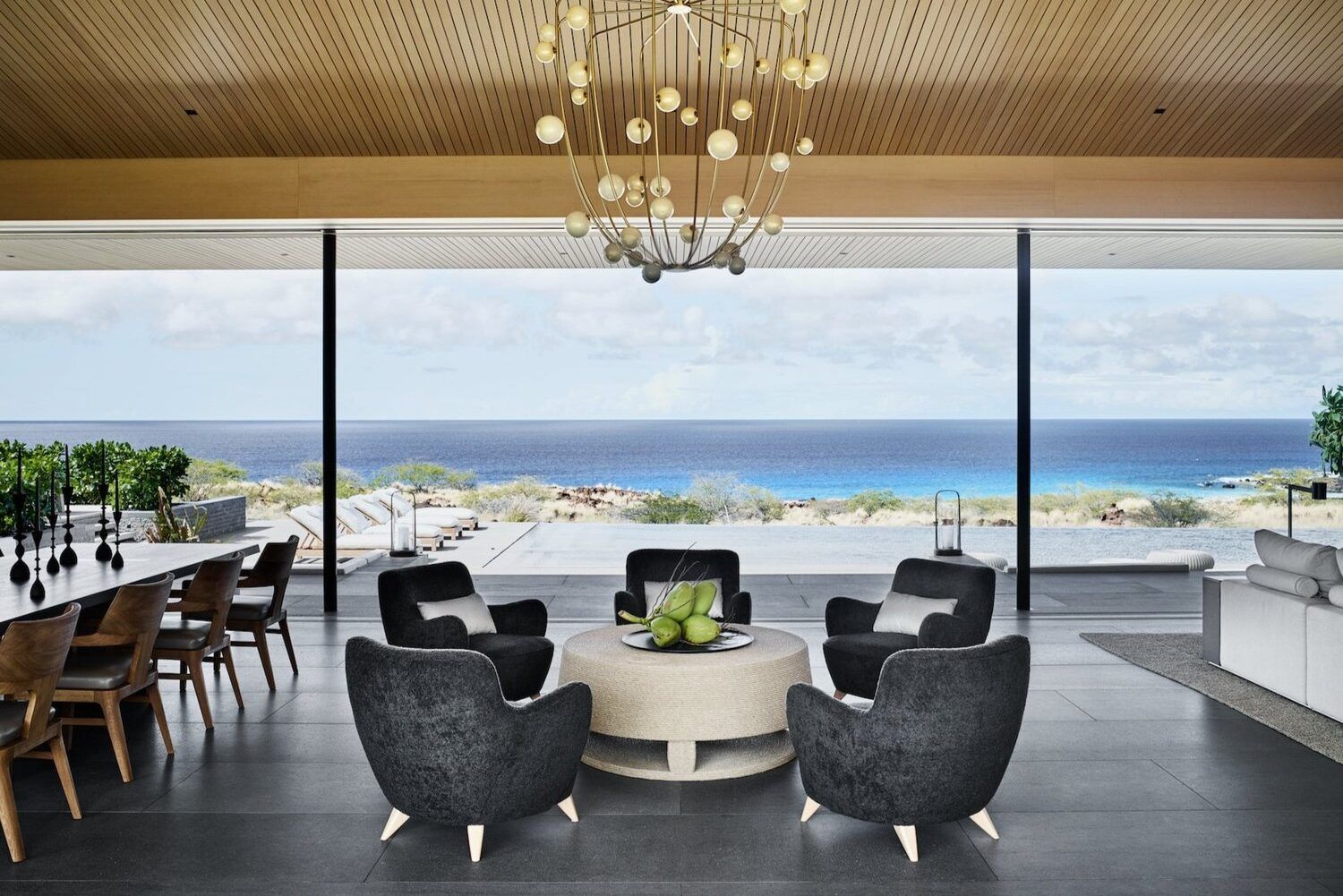 View of open concept upscale living area with table and chairs and a cozy seating area in foreground. In background is a glass wall overlooking the pool area and beyond the ocean.