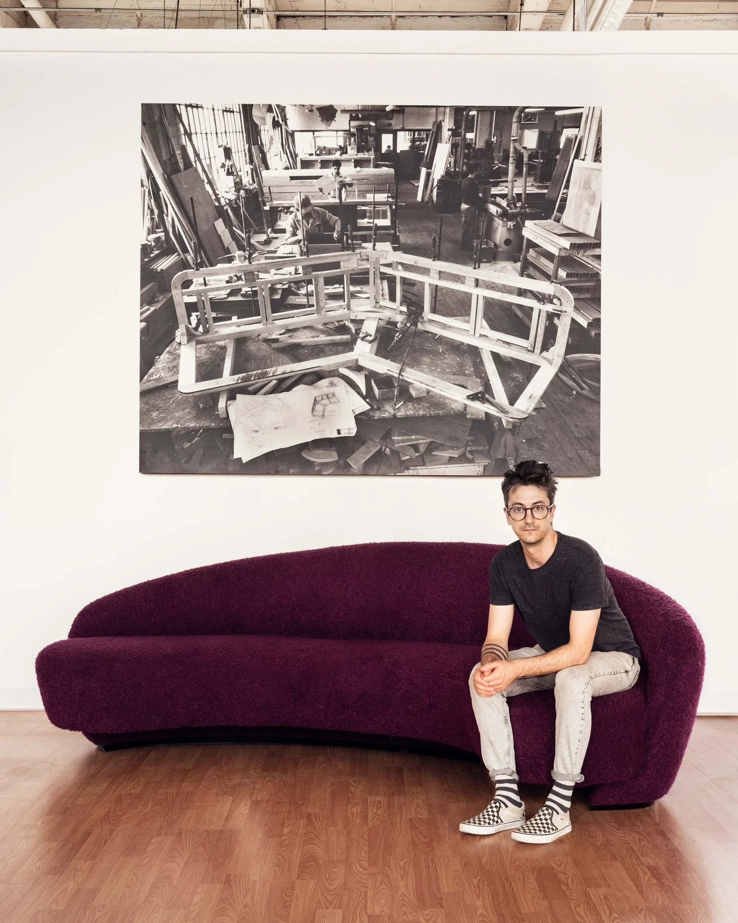 Casually dressed young man looking at camera while sitting on a small stylized grape color couch. Hanging on wall behind is a sepia-toned vintage image of a furniture maker at work in his studio.