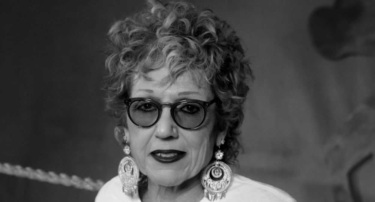 Black and white image of stylish woman in her 60's wearing oversized accessories, jeans and a t-shirt that says "Judy Chicago".