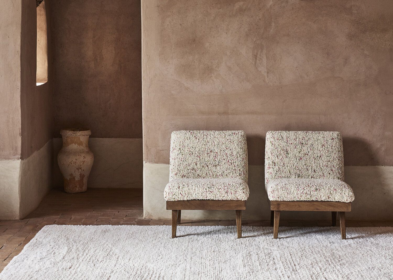 Adobe style walls with two wood based simple chairs upholstered with slubby boucle fabric on a woven rug.