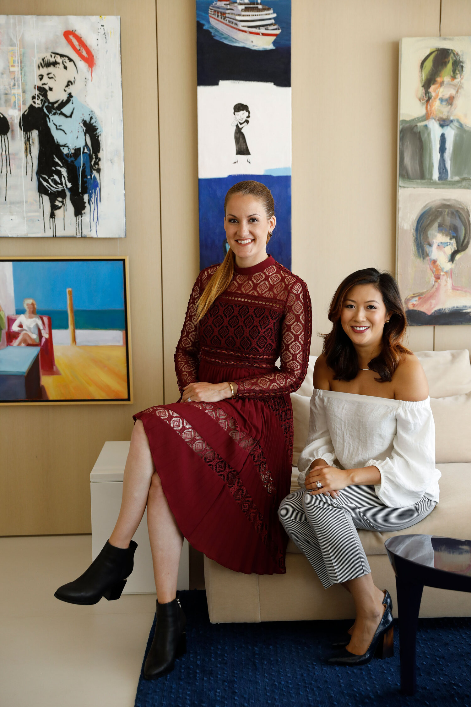 Two young women seated and smiling for camera with modern artwork in background.