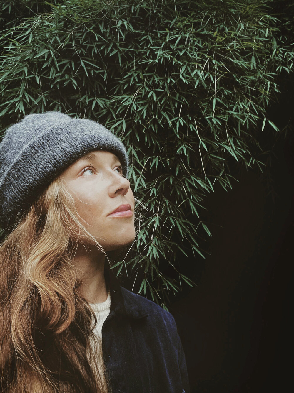 Young woman with long hair in knit cap looking up at a large green bush.