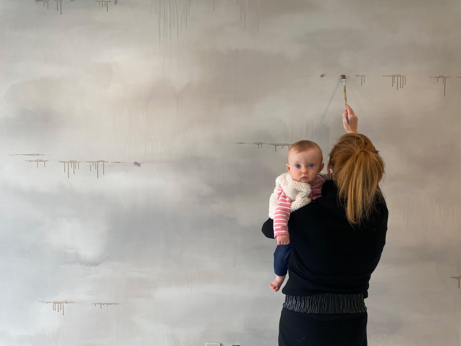 Woman holding small baby while painting a wall.