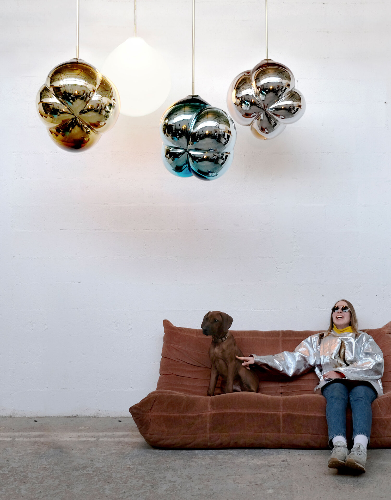 Four pendulum lights, one a white sphere, the other three are different metallic toned organic shapes like balloons over a laughing woman seated on a couch with a dog.
