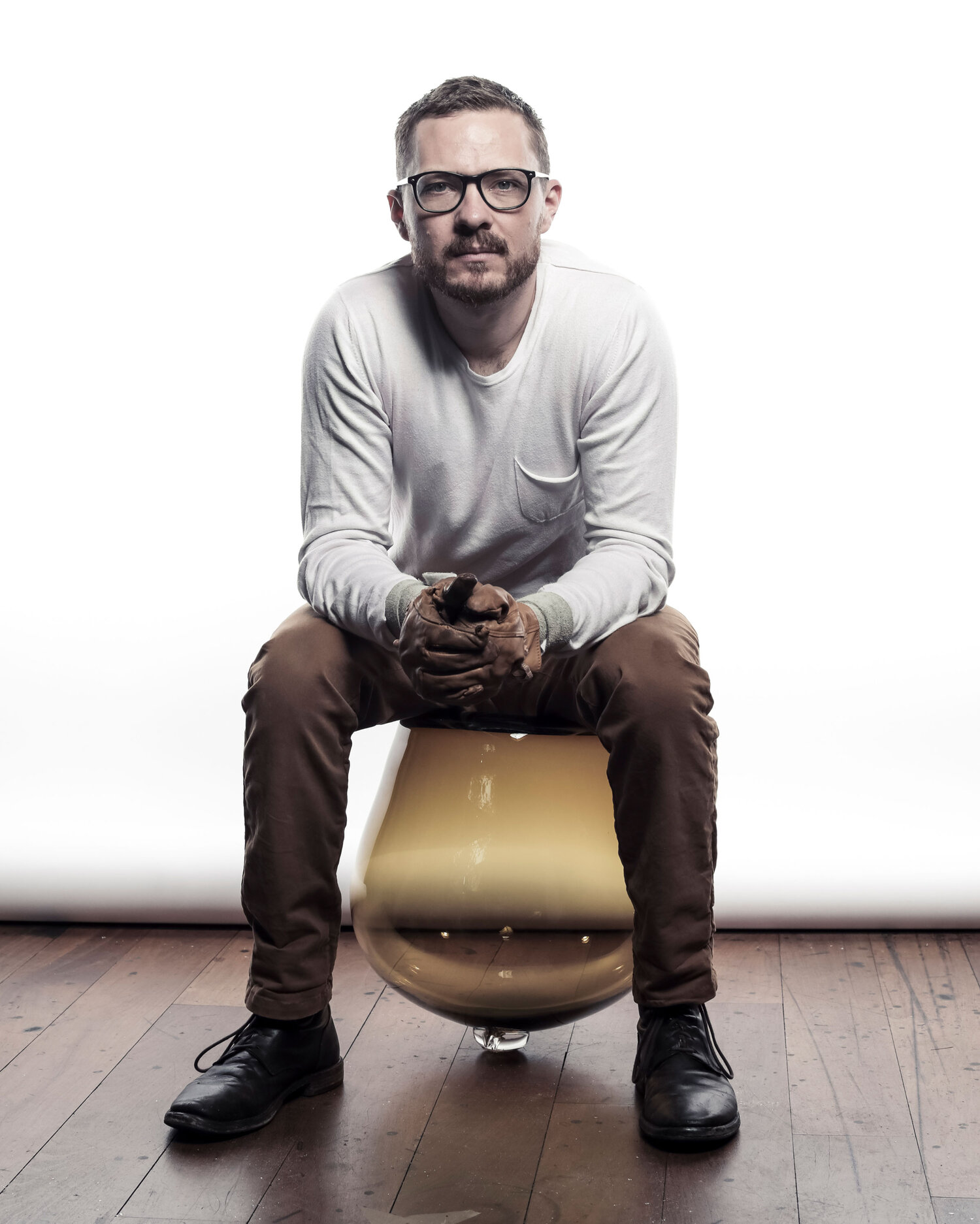 Man in glasses perched on a glass modern stool while he looks at camera.
